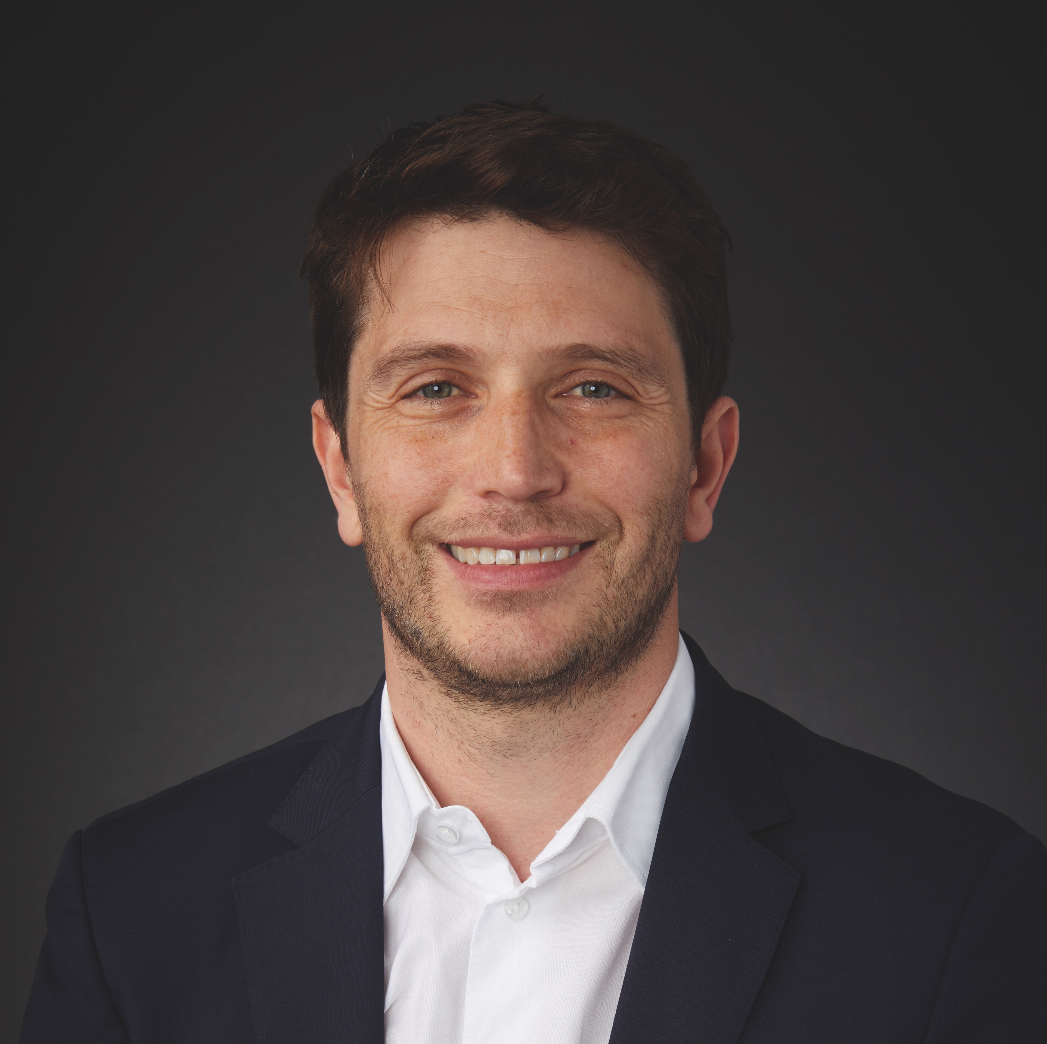 A man with short brown hair and a trimmed beard, wearing a white shirt and dark blazer, smiles at the camera against a plain dark background.