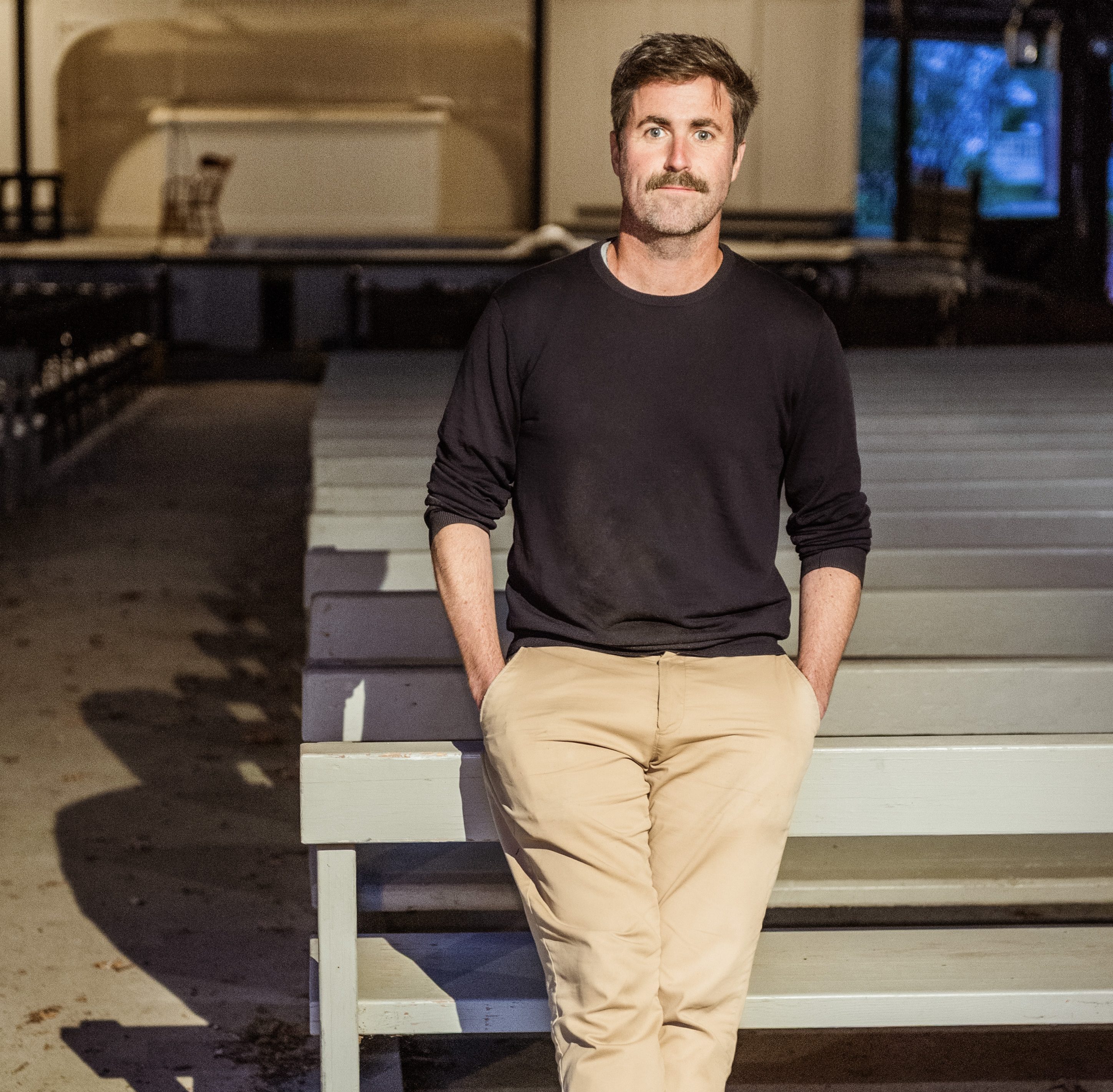 A man with a mustache, wearing a black shirt and beige pants, stands with hands in pockets in front of empty wooden benches in an indoor space.