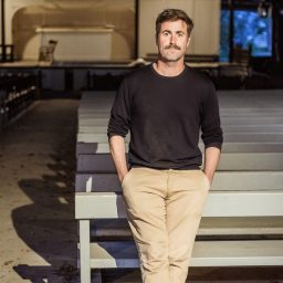 A man with a mustache, wearing a black shirt and beige pants, stands with hands in pockets in front of empty wooden benches in an indoor space.