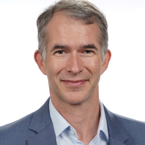 A middle-aged man with short gray hair, wearing a blue blazer and light blue shirt, smiling at the camera against a plain white background.