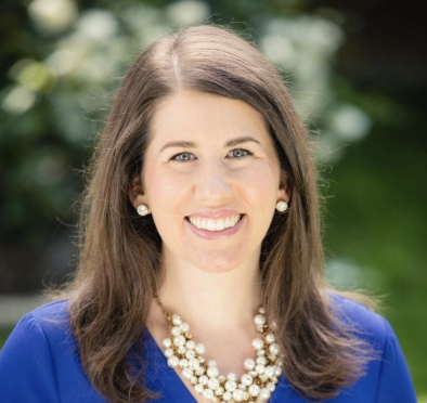 Woman with long brown hair, wearing a blue top, pearl necklace, and earrings, smiling outdoors with greenery in the background.