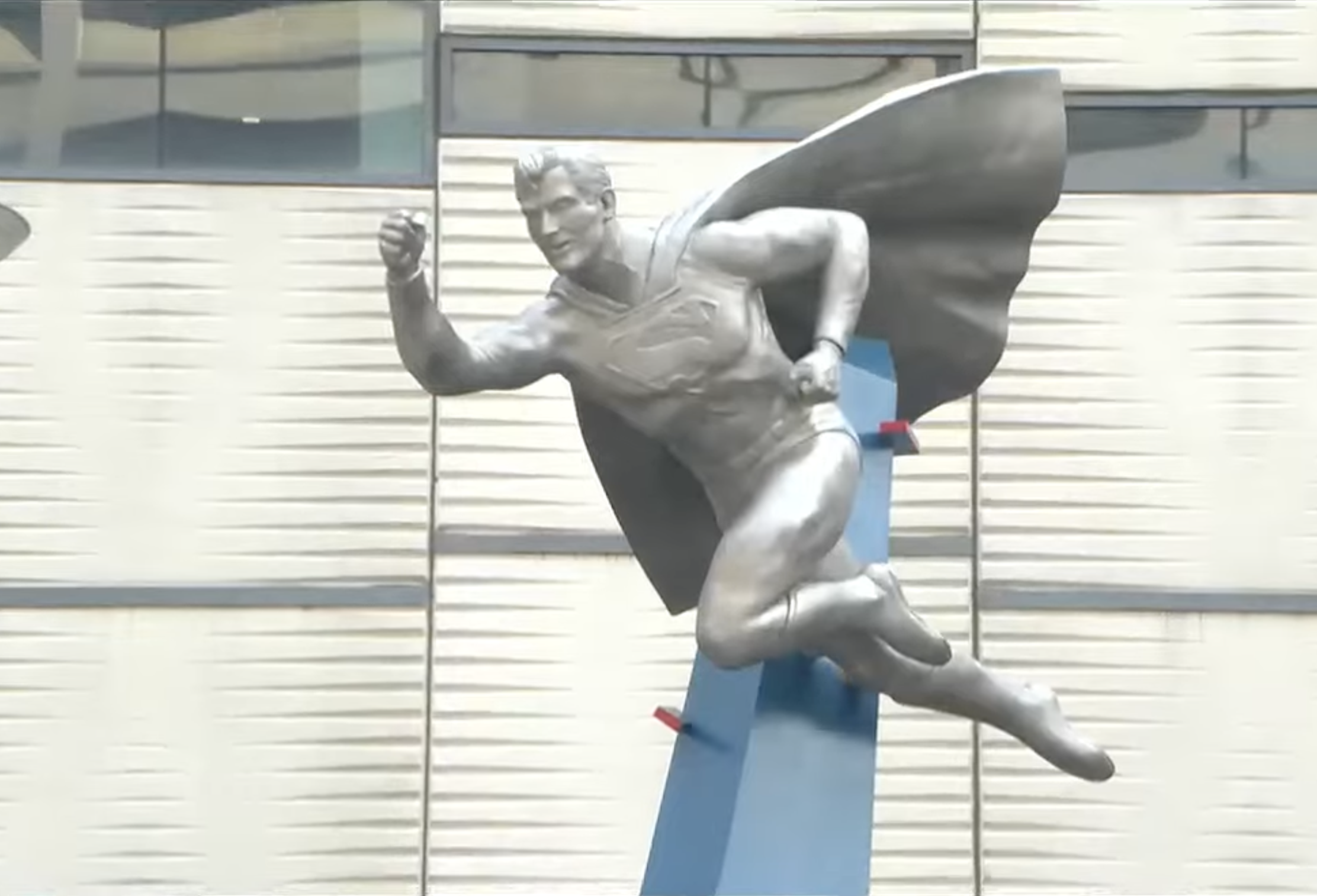 A metallic statue of a superhero with a cape, posed as if flying, mounted on a blue pedestal in front of a modern building.