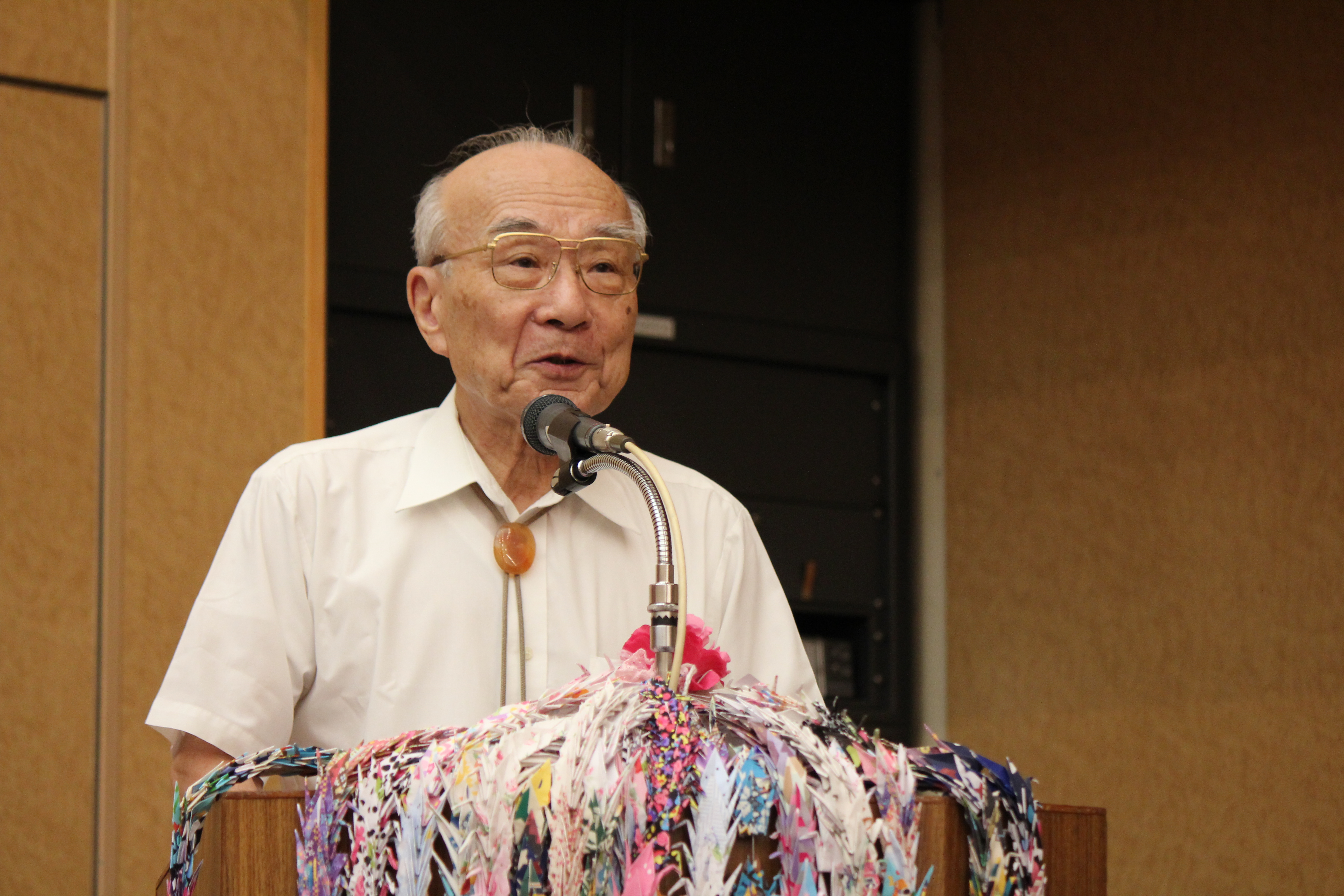 An elderly man in a white shirt speaks at a podium adorned with colorful folded paper cranes and a microphone in an indoor setting.
