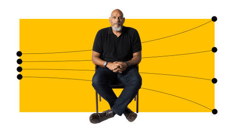 A man with a bald head and beard sits on a chair with arms resting on his lap, in front of a yellow rectangle with black lines and dots extending outward.