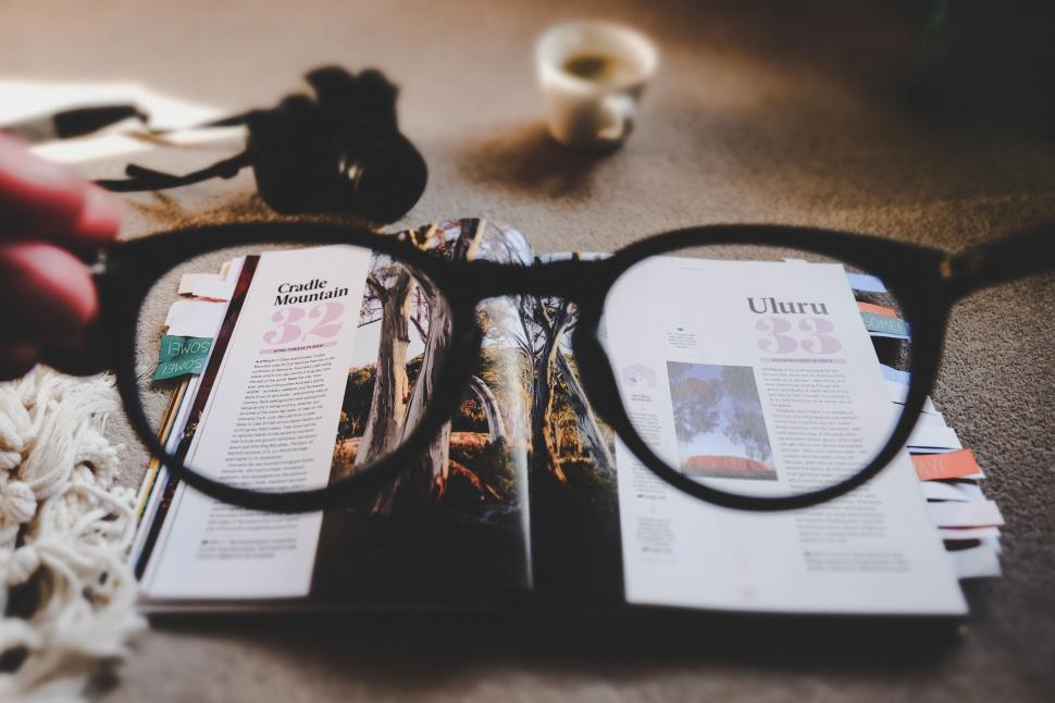 A pair of eyeglasses held in front of an open magazine with articles and photos, hinting at the science of LASIK, with a blurred camera and a cup in the background.