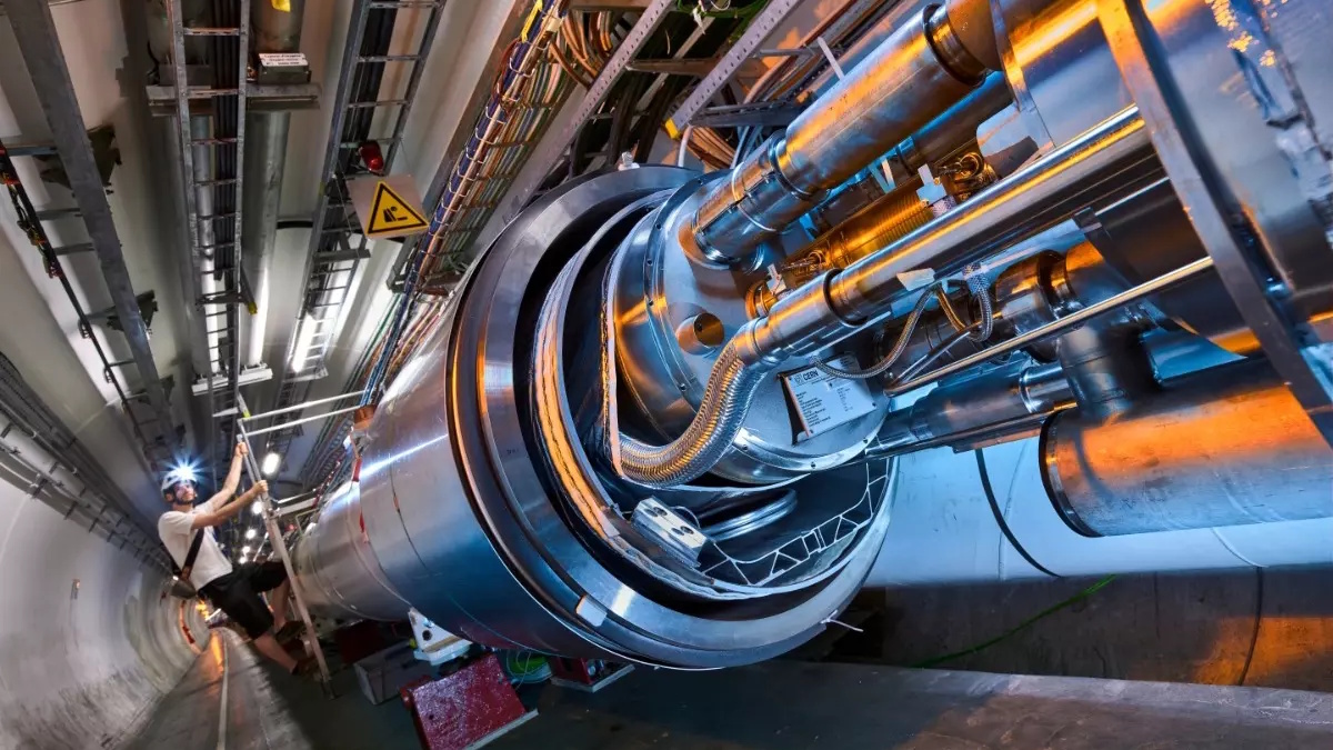 A person inspects a large, cylindrical section of a Higgs factory tunnel lined with metal pipes, cables, and equipment—a crucial site for particle physics research.