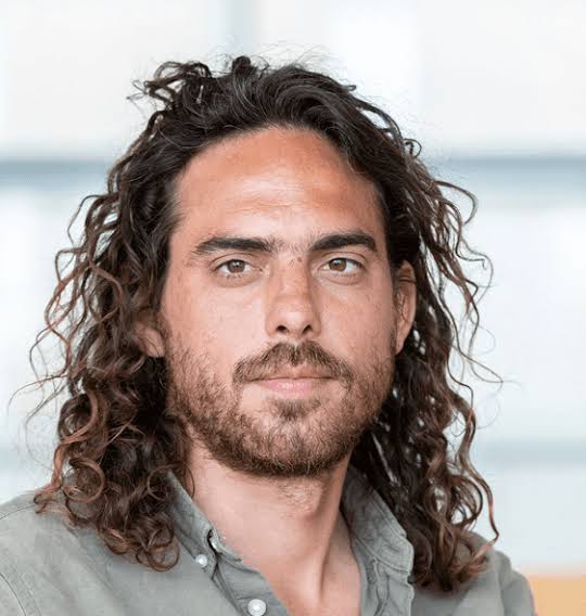 A man with long curly hair and a beard looks directly at the camera, wearing a button-up shirt with a neutral background.
