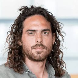 A man with long curly hair and a beard looks directly at the camera, wearing a button-up shirt with a neutral background.