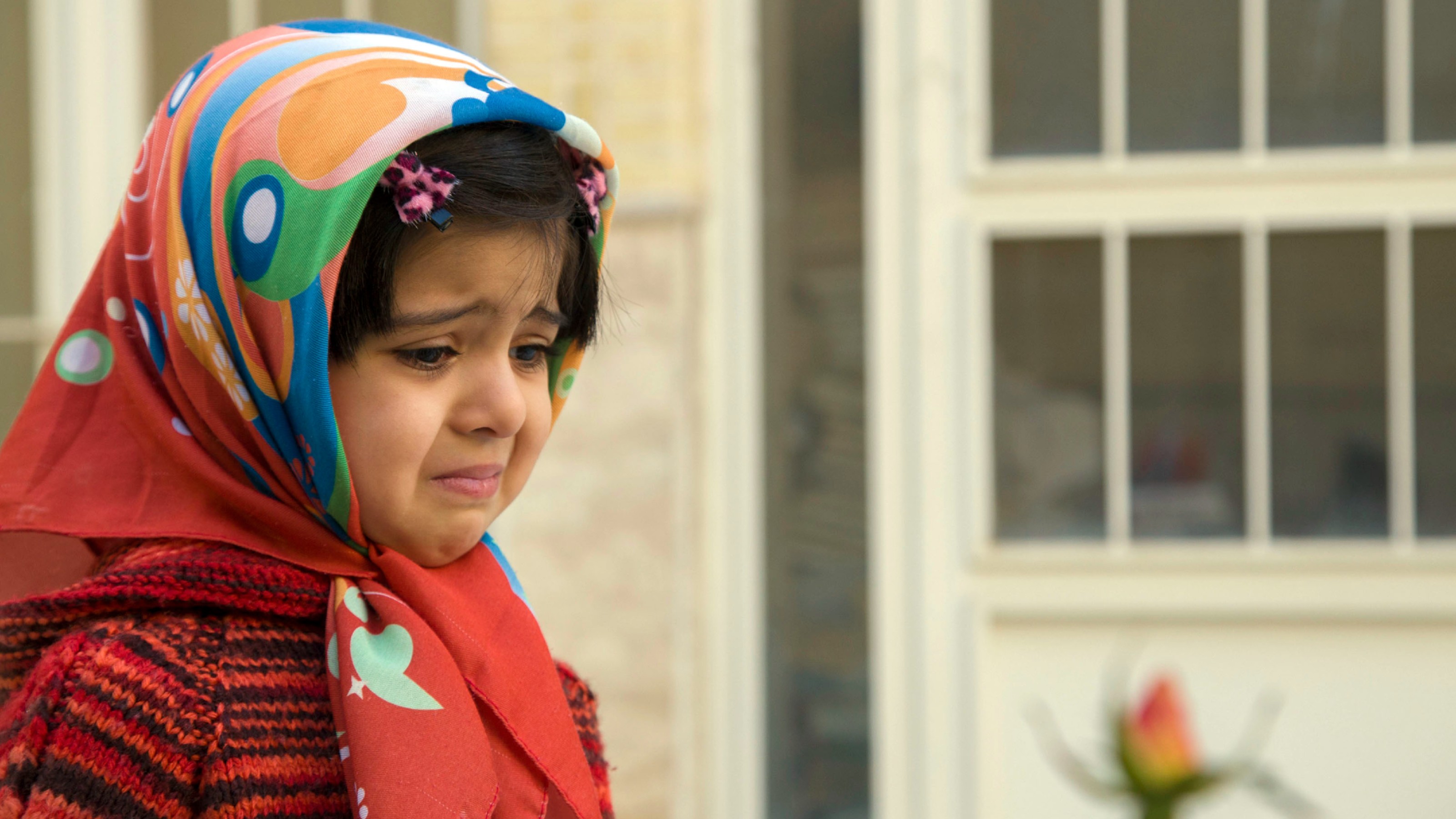 A young girl wearing a colorful headscarf and red sweater stands outside, looking sad with a slight frown. A blurred flower appears in the foreground.