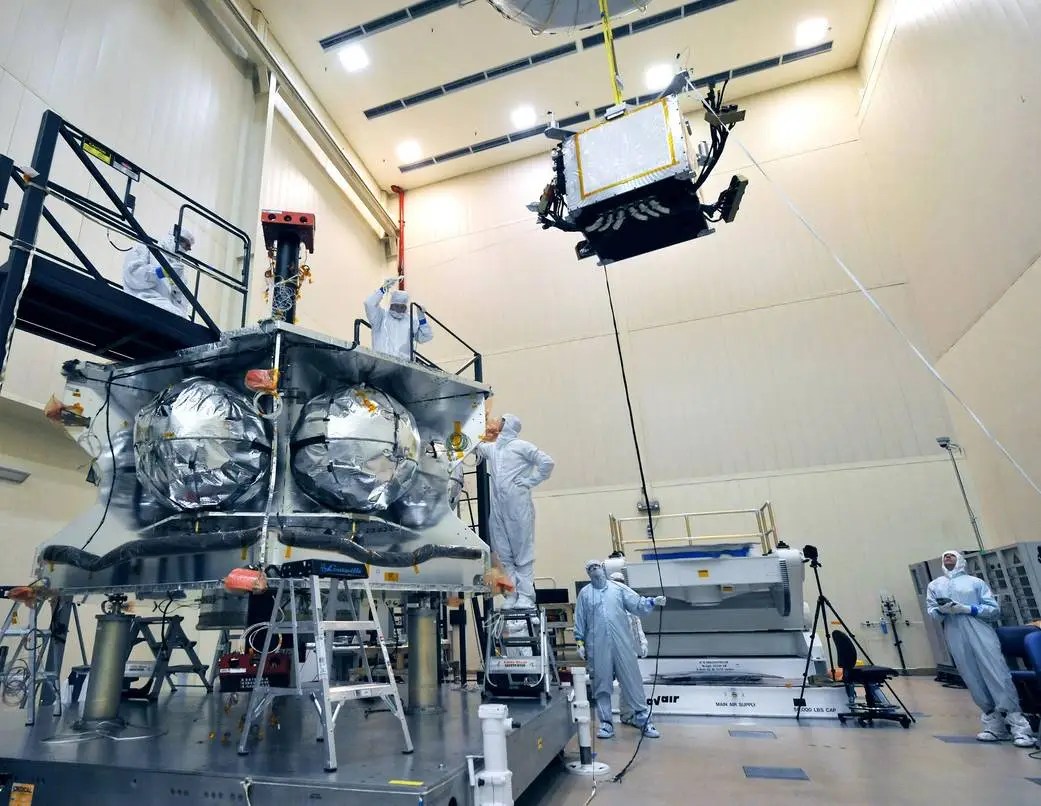 Technicians in protective suits work in a clean room, guiding a satellite component—part of NASA's Juno mission—being lowered by a crane onto a larger spacecraft structure.