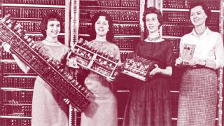 Four women stand in front of a vintage computer, each holding different components of its hardware, with the computer's panel visible in the background—an image reminiscent of historical innovation studied by Angus Fletcher.