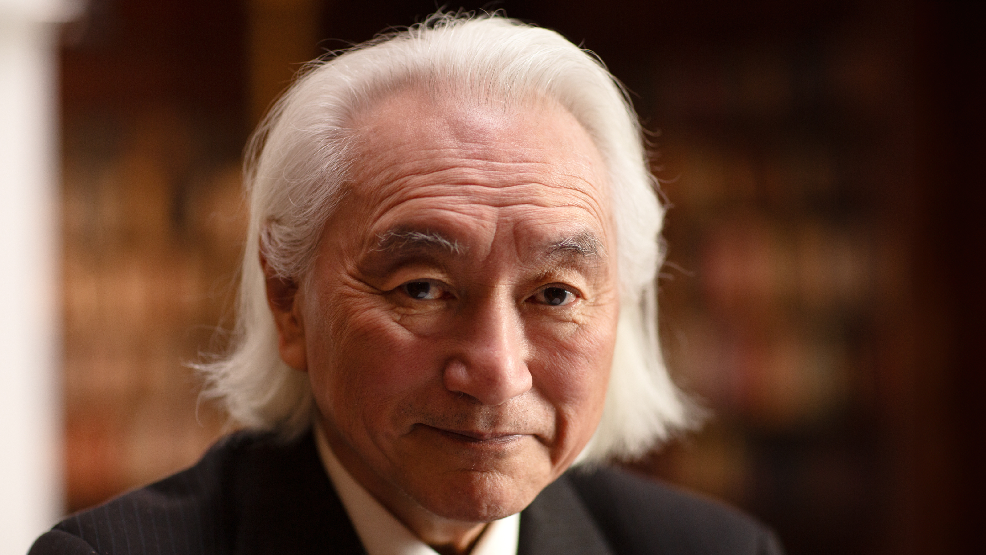 An older man with long white hair and a suit looks at the camera, standing in front of a blurred background with bookshelves.