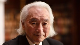 An older man with long white hair and a suit looks at the camera, standing in front of a blurred background with bookshelves.