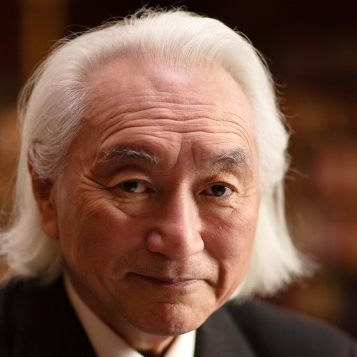 An older man with long white hair and a suit looks at the camera, standing in front of a blurred background with bookshelves.
