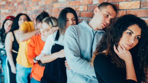 A group of seven young adults stands in a line against a brick wall, each appearing bored or tired, with some resting their heads on others' shoulders.