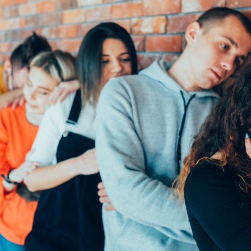 A group of seven young adults stands in a line against a brick wall, each appearing bored or tired, with some resting their heads on others' shoulders.