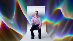 A man sits on a chair in front of a white backdrop with vibrant, rainbow-colored abstract patterns in the background.
