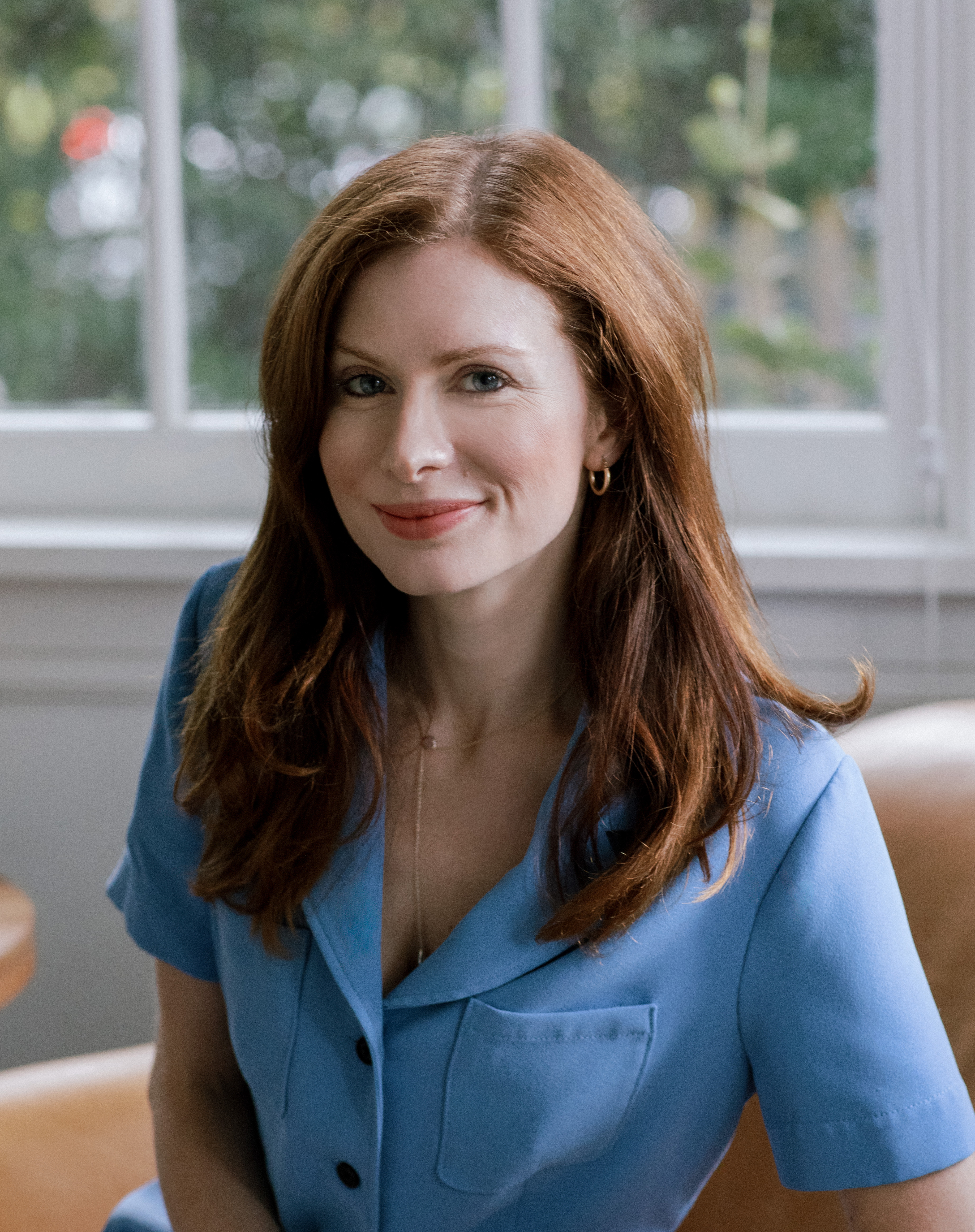 A woman with long red hair wearing a blue button-up shirt sits on a tan chair in front of a window with greenery outside.