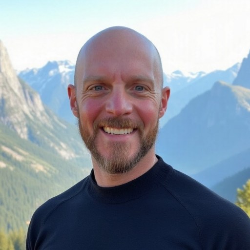 A smiling bald man with a trimmed beard stands outdoors, with mountains and a clear sky in the background.