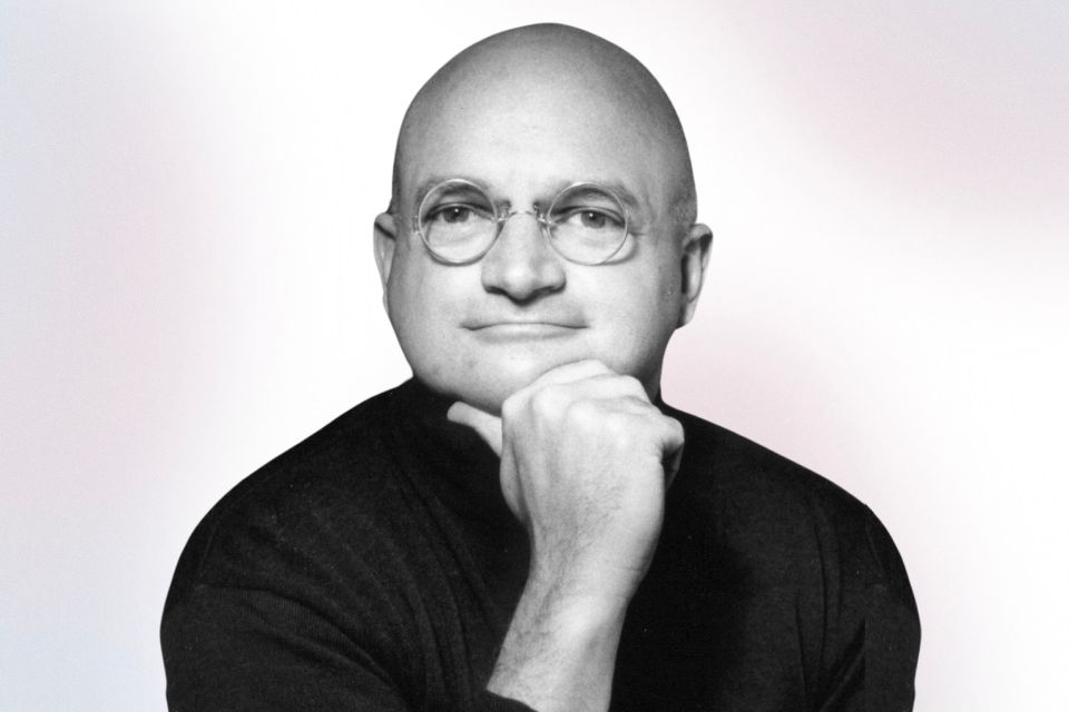 A bald man wearing round glasses and a dark shirt rests his chin on his hand, looking at the camera against a light background.