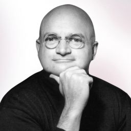 A bald man wearing round glasses and a dark shirt rests his chin on his hand, looking at the camera against a light background.