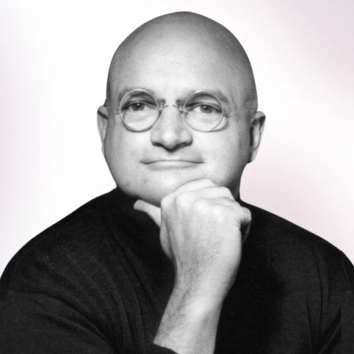 A bald man wearing round glasses and a dark shirt rests his chin on his hand, looking at the camera against a light background.