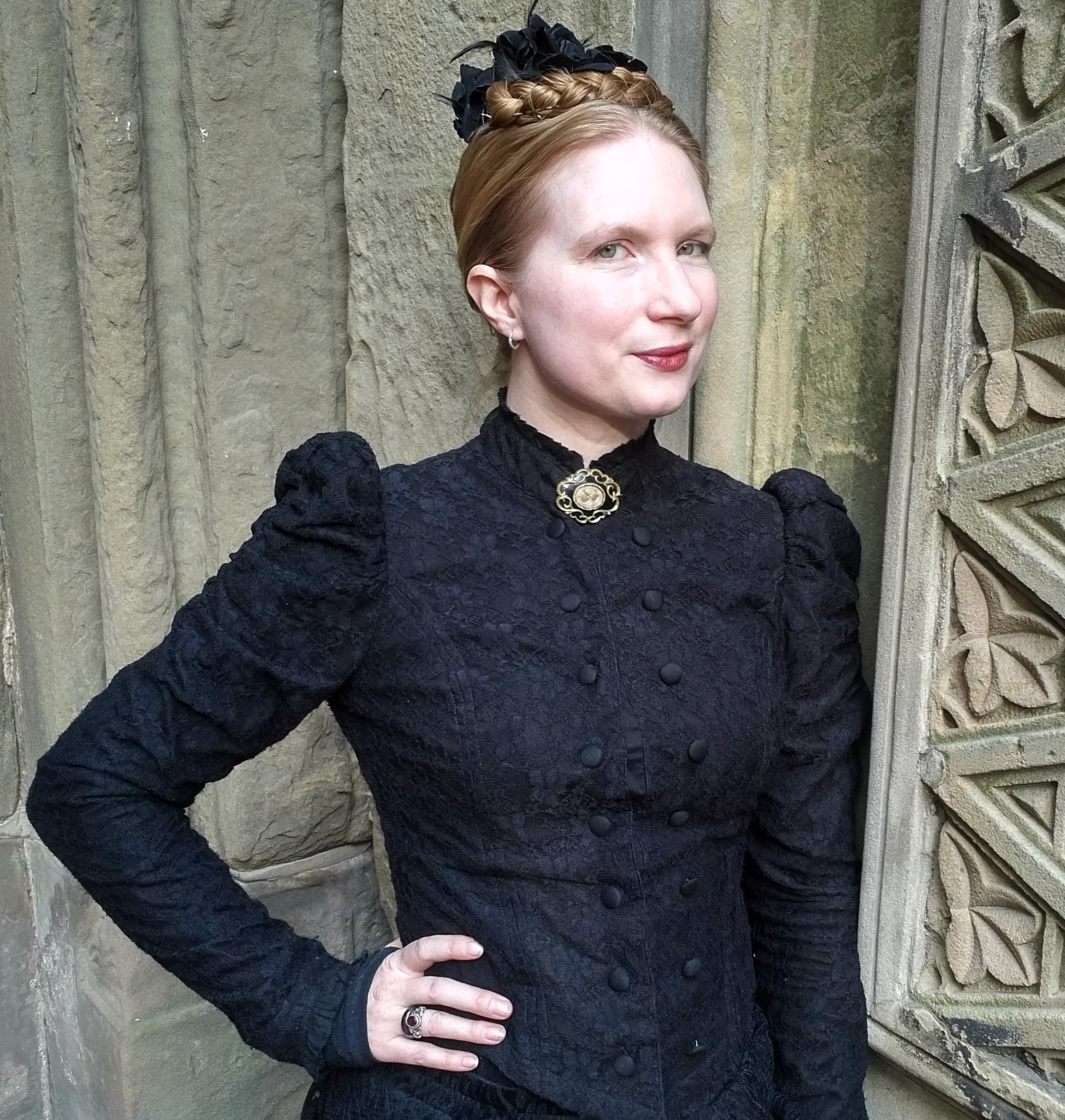 A woman wearing a high-necked, black Victorian-style dress poses near a stone wall with architectural details.
