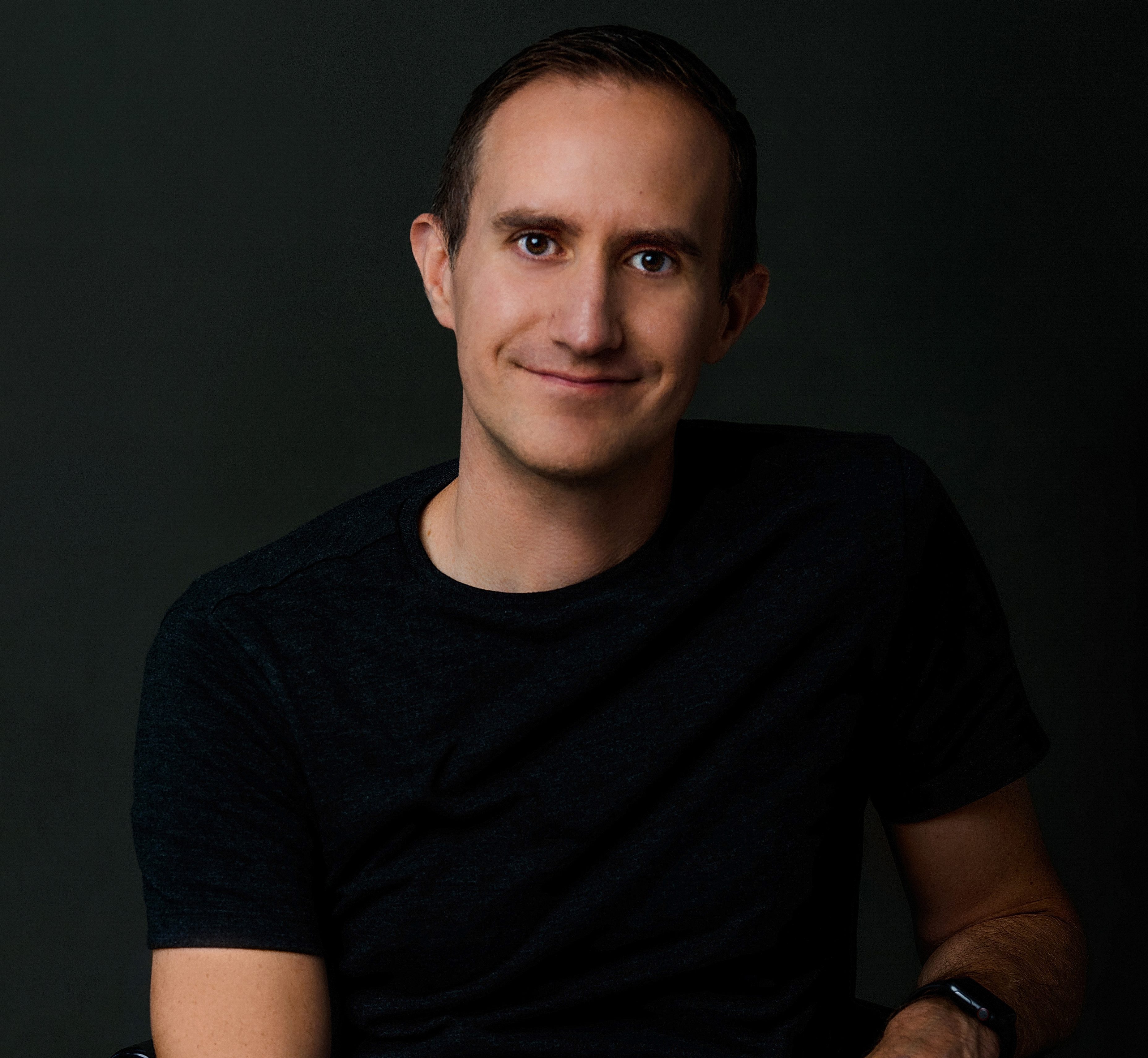 A man in a dark t-shirt sits against a dark background, smiling with his hands clasped in his lap.