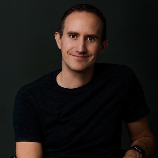 A man in a dark t-shirt sits against a dark background, smiling with his hands clasped in his lap.