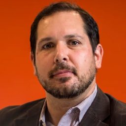 A man with short dark hair and a beard, wearing a checked shirt and dark blazer, poses in front of an orange background.