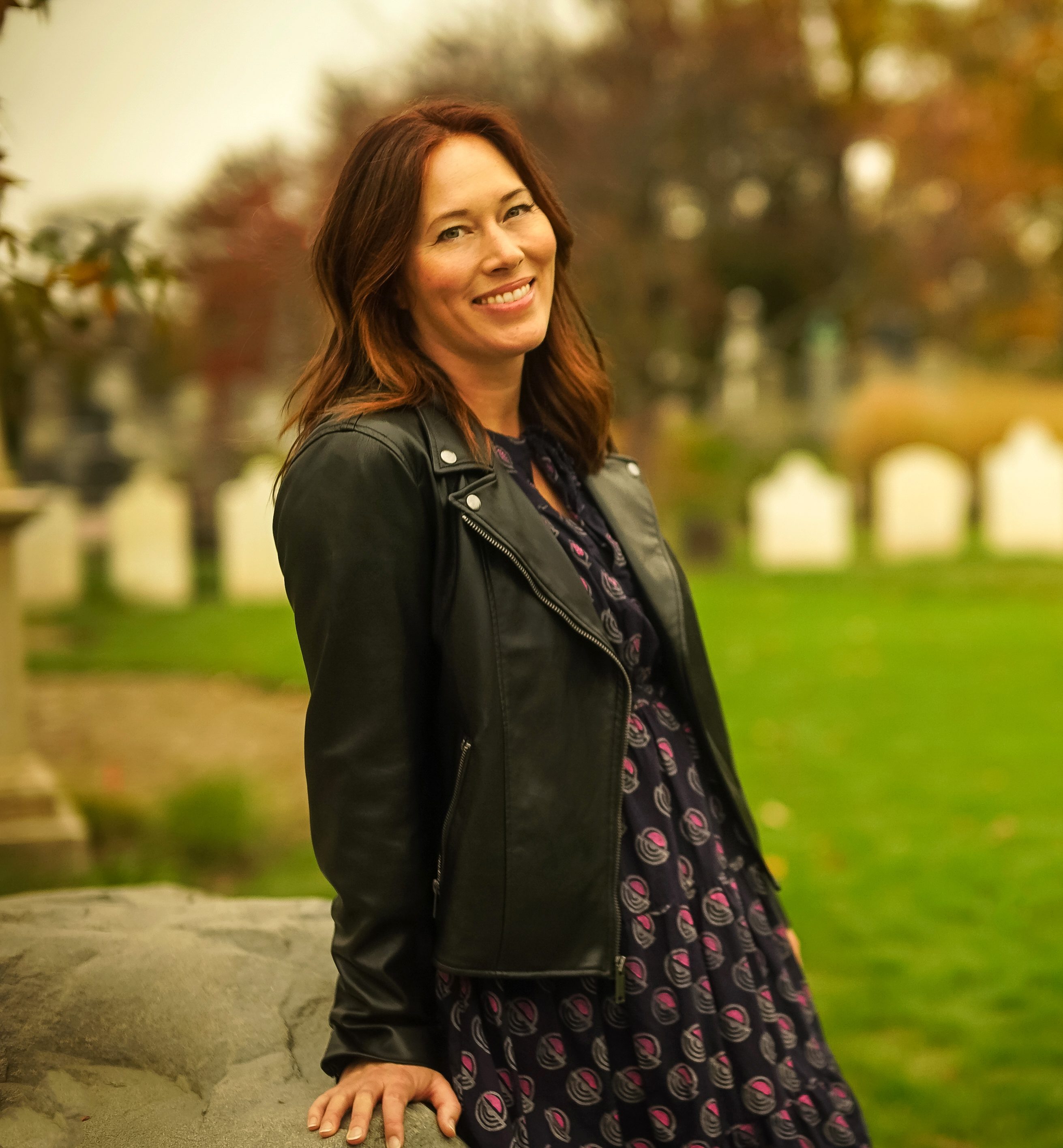 A woman in a patterned dress and black jacket stands outdoors near a stone, smiling, with green grass and gravestones visible in the background.