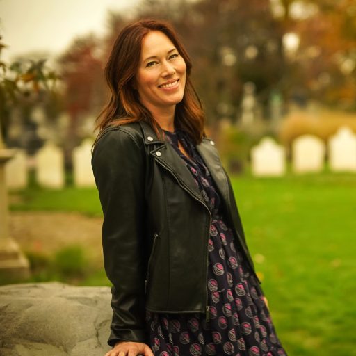 A woman in a patterned dress and black jacket stands outdoors near a stone, smiling, with green grass and gravestones visible in the background.