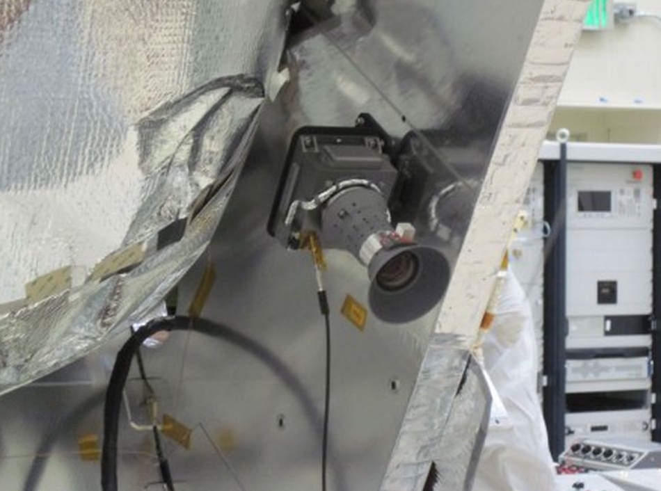 A close-up view of a camera mounted on a metallic, foil-covered structure, with wires and equipment visible nearby during NASA's Juno mission.
