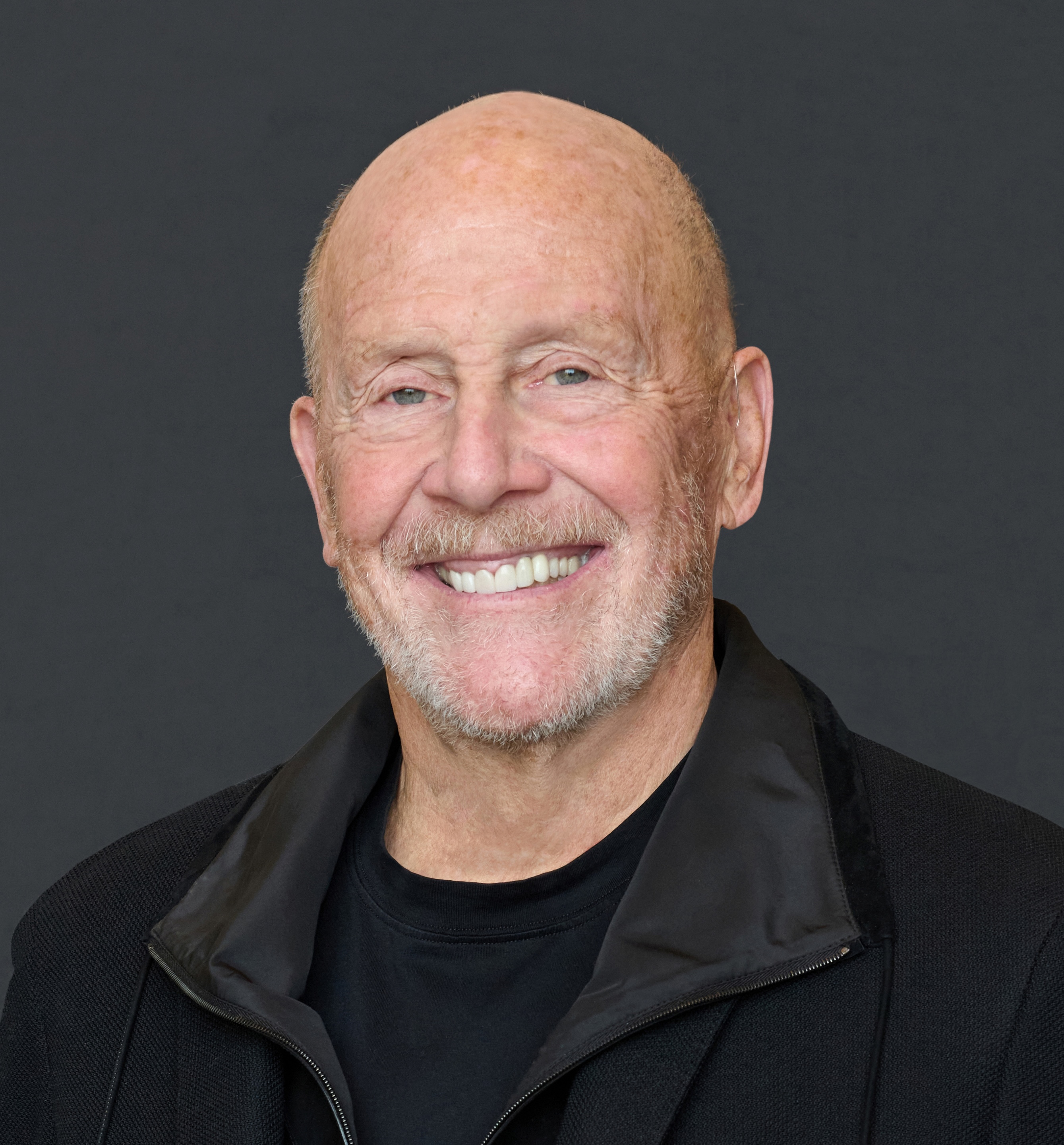 A smiling older man with a bald head and trimmed white beard, wearing a black jacket and black shirt, in front of a plain dark background.
