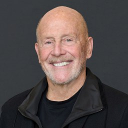 A smiling older man with a bald head and trimmed white beard, wearing a black jacket and black shirt, in front of a plain dark background.
