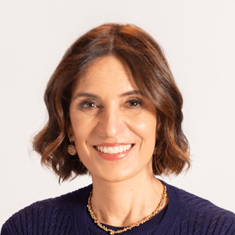 A woman with shoulder-length brown hair, wearing a navy sweater, gold necklace, and earrings, smiles at the camera against a plain light background.