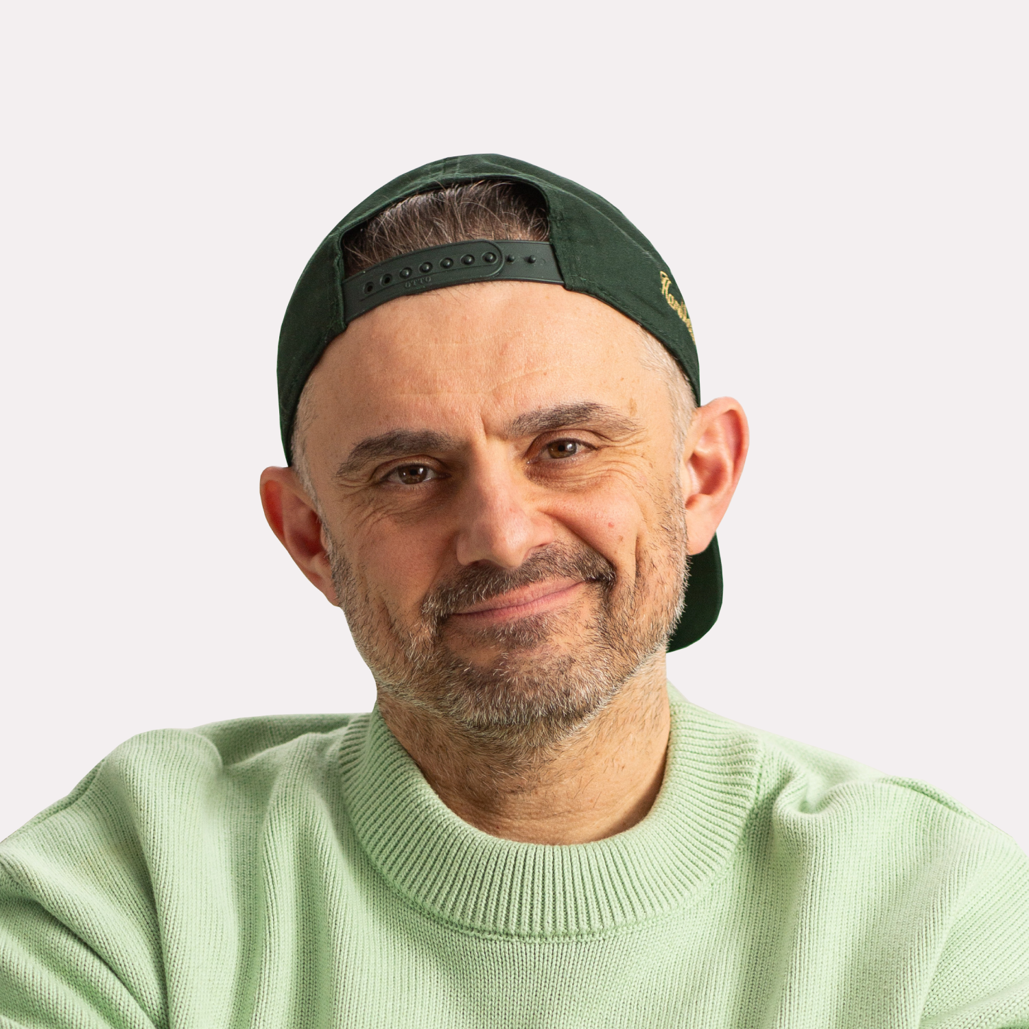 A man with a beard wearing a light green sweater and a dark green baseball cap backward, smiling with a neutral background.