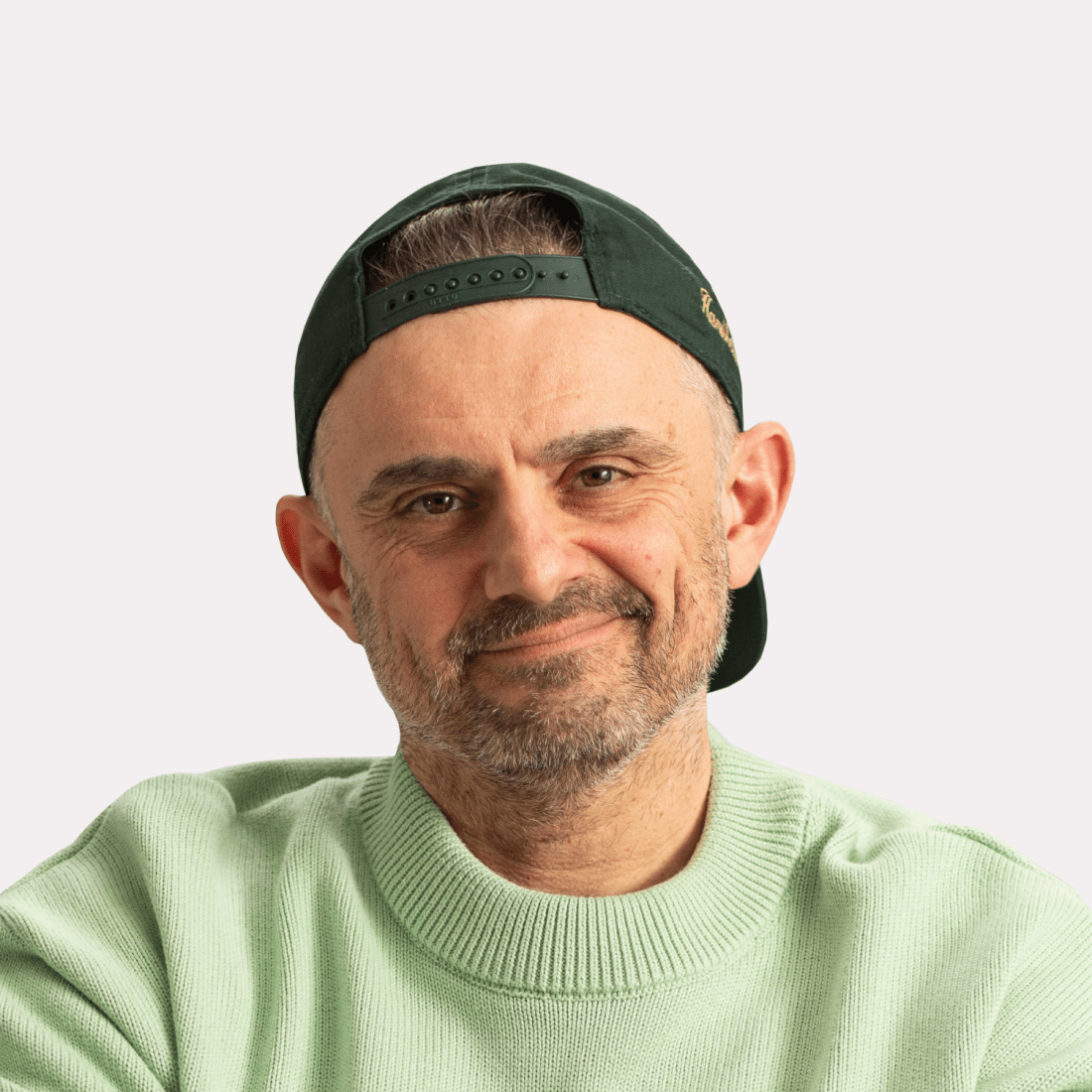 A man with a beard wearing a light green sweater and a dark green baseball cap backward, smiling with a neutral background.