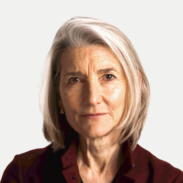 Portrait of an older woman with gray hair, parted to the side, wearing a dark top, looking directly at the camera with a neutral expression against a plain background.