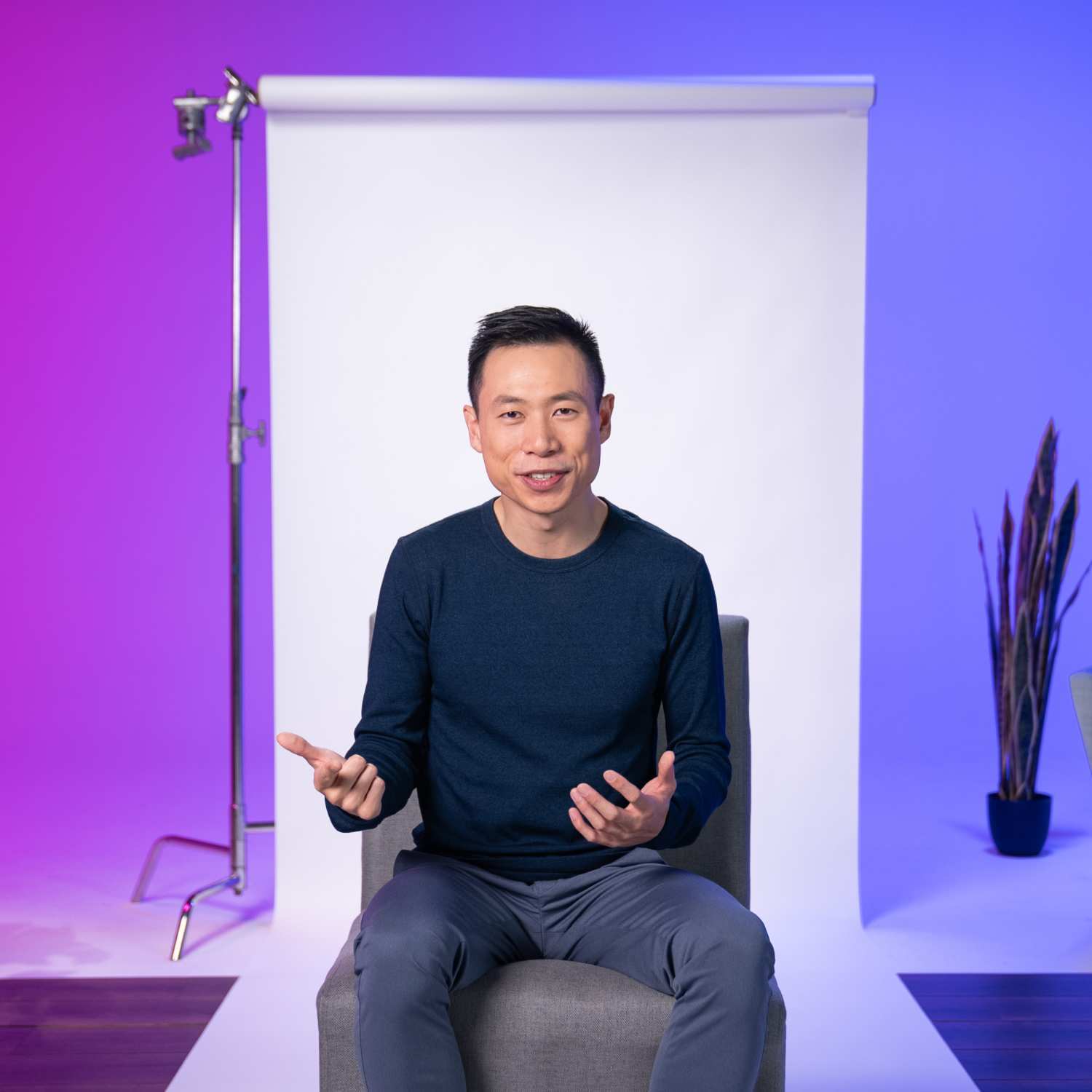 A person sits on a chair in front of a white backdrop, gesturing with both hands. The background is lit with purple and blue tones, and a plant and lighting equipment are visible.