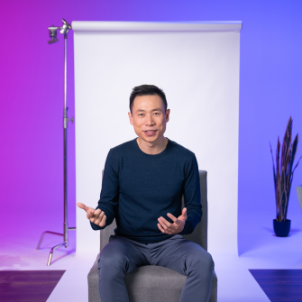 A person sits on a chair in front of a white backdrop, gesturing with both hands. The background is lit with purple and blue tones, and a plant and lighting equipment are visible.
