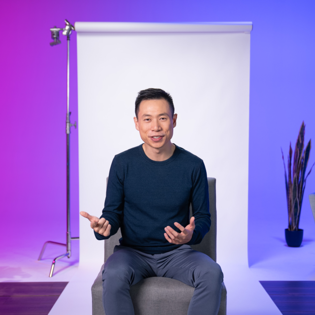 A person sits on a chair in front of a white backdrop, gesturing with both hands. The background is lit with purple and blue tones, and a plant and lighting equipment are visible.
