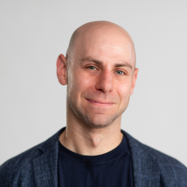A bald man wearing a dark blazer and black shirt smiles slightly against a plain light gray background.