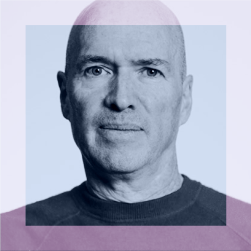 A middle-aged bald man with light skin and blue eyes, wearing a dark crewneck shirt, looks directly at the camera against a plain background.