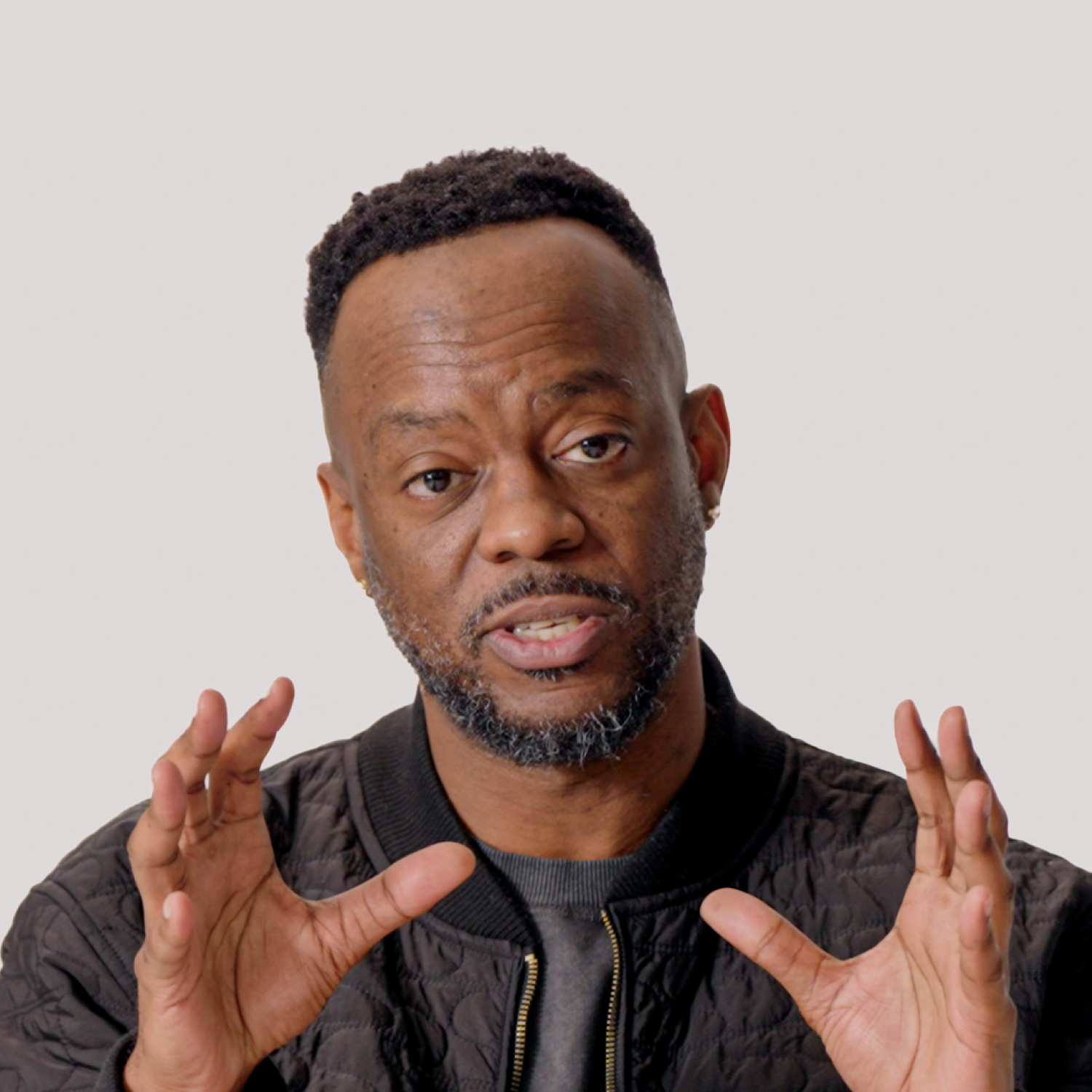 A man with short hair and a beard speaks while gesturing with both hands; he wears a black quilted jacket against a plain light background.