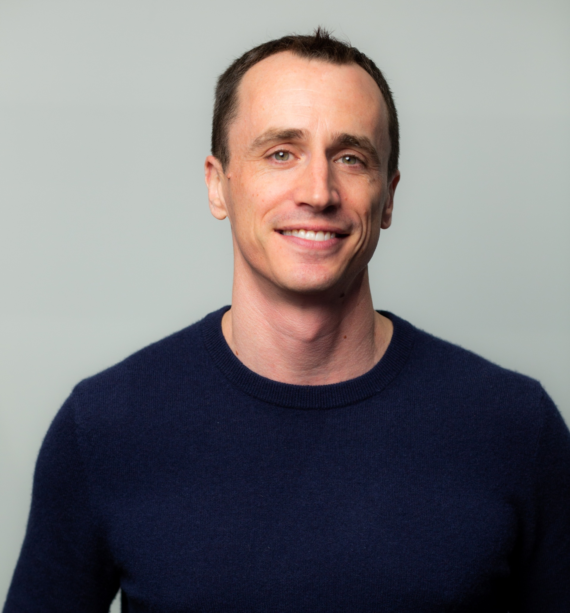 A man with short dark hair wearing a navy blue sweater smiles at the camera against a plain light gray background.