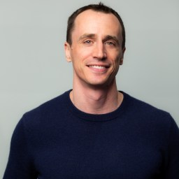 A man with short dark hair wearing a navy blue sweater smiles at the camera against a plain light gray background.