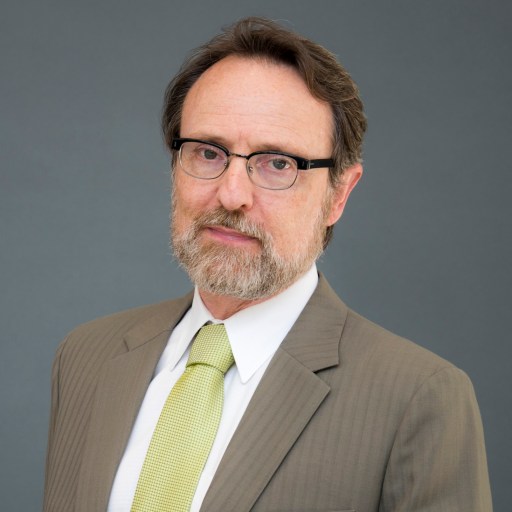 A middle-aged man with glasses, a beard, and short brown hair wears a tan suit, white shirt, and light green tie against a plain gray background.