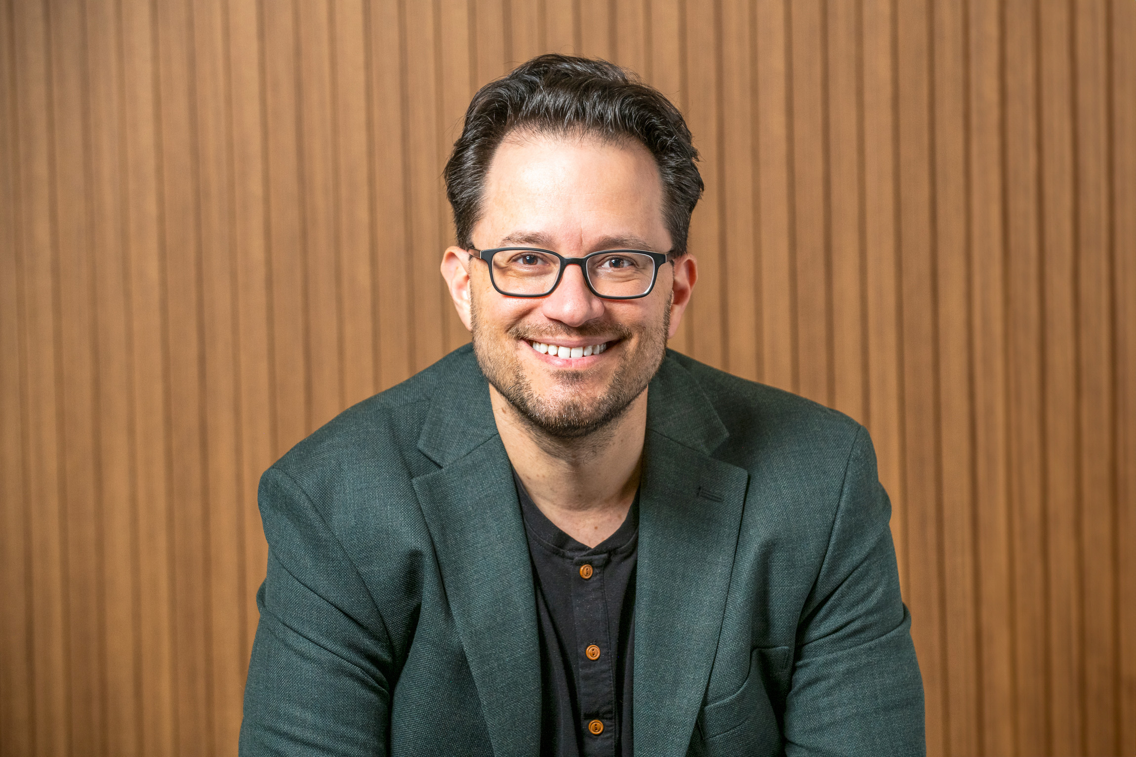 A man with glasses and a beard, wearing a dark green blazer and black shirt, smiles while seated in front of a wooden slat background.