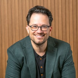 A man with glasses and a beard, wearing a dark green blazer and black shirt, smiles while seated in front of a wooden slat background.